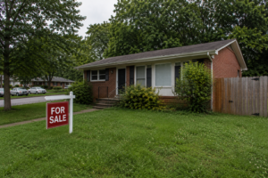 A modest 1970s ranch home in a Raleigh NC neighborhood on an overcast afternoon, showing weathered siding, an overgrown shrub line, and a slightly faded For Sale sign in the front yard