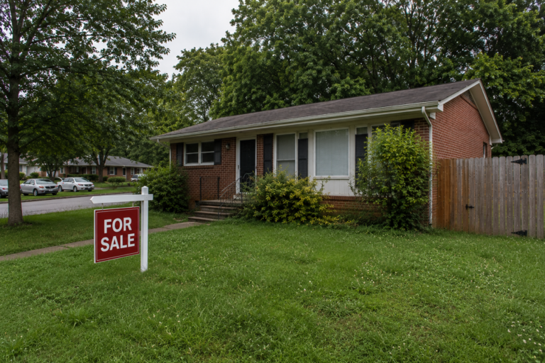 A modest 1970s ranch home in a Raleigh NC neighborhood on an overcast afternoon, showing weathered siding, an overgrown shrub line, and a slightly faded For Sale sign in the front yard