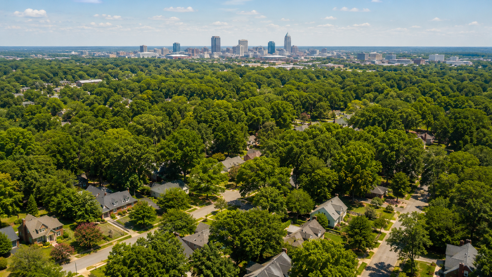 Aerial view of Raleigh, NC residential neighborhoods in summer 2026 with the downtown skyline visible in the distance, illustrating continued growth in the Wake County real estate market.
